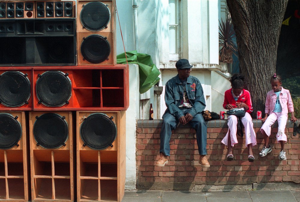 Description – Family sitting on a wall beside a soundsystem at Notting Hill Carnival 2014