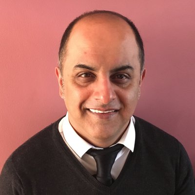 A picture of Bali Rai - a middle-aged Brown man with a baling head who is smiling in front of a pink background