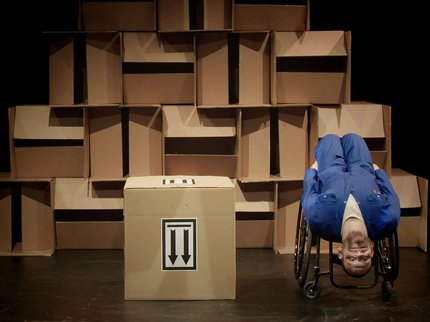 Daryl, a white man wearing a blue overall sits upside down in his wheelchair, his head almost resting on the floor as he looks outwards. Next to him is a large cardboard box with 2 arrows on it pointing to the floor. The background is a wall of boxes