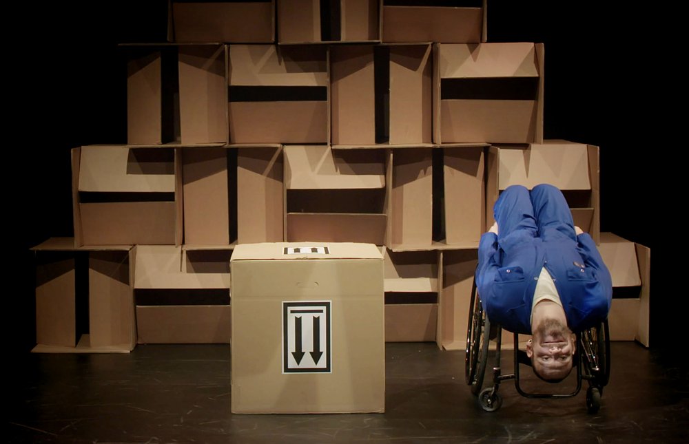 A man in blue overalls  sits upside down on a wheelchair with brown cardboard boxes in the background.