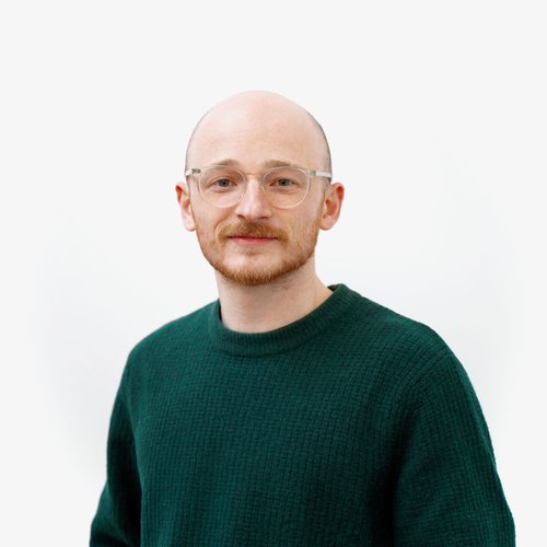 Male with clear framed glasses, short ginger facial hair smiling to camera. Wearing a green knitted jumper, standing against a white background.