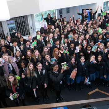 Author Sufiya Ahmed wears a black suit and stands next to the ELS banner in a school hall