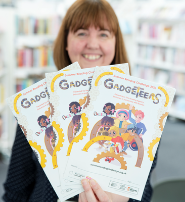Smiling, dark haired female, holds up 3 illustrated Gadgeteers collector posters