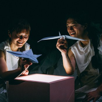 Two people holding origami in front of a cardboard box.