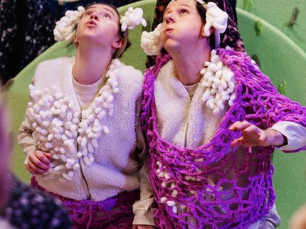 Two performers dressed as sheep, with white woolly costumes and curly headpieces, are kneeling and looking up in awe at glowing lights held above them by a third performer. The scene appears whimsical and theatrical. Bold yellow text reads “Counting