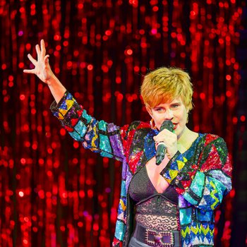 A woman with short hair wearing a colourful jacket raising one hand and speaking into a microphone.