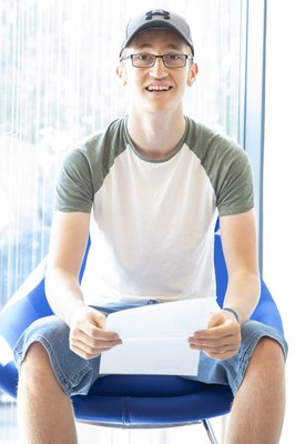 A young man in a hat and glasses sitting in a blue chair holds his GCSE results and smiles.