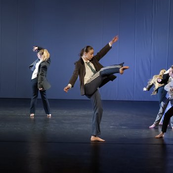 Six young dancers on stage. The front dancer is kicking her right leg in to the air.