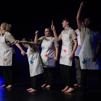 Five dancers on stage wearing paint splattered aprons and holding paintbrushes.