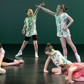 Seven children dancing on stage wearing pastel coloured t shirts. Two are pointing to the sky.