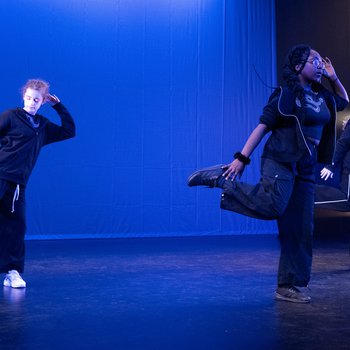 Three dancers on stage wearing black outfits, raising one leg.