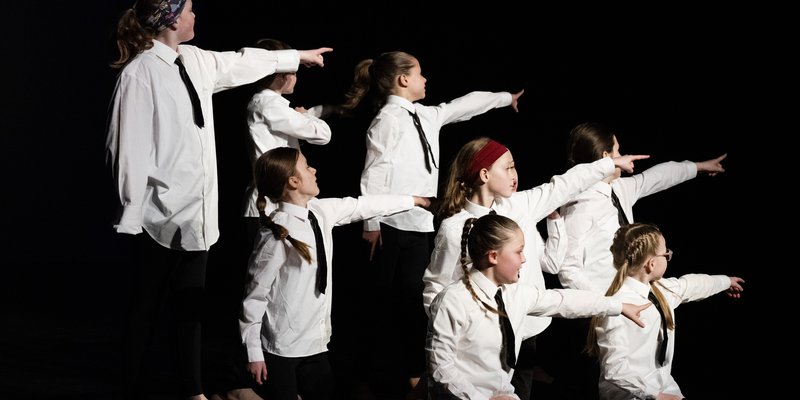 8 young people wearing white shirts and black ties on stage pointing to the right