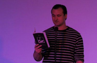 Photograph of Poet Chris Lanyon, a white man with short dark hair, reading from a book into a microphone