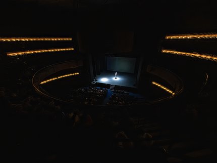 A dark image of a theatre. One person is performing on stage.