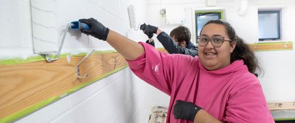 An Inspire College learner in a pink hoodie painting a wall.