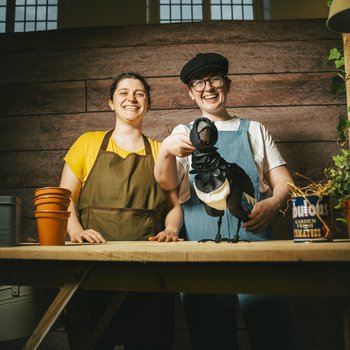 Three characters form Along Came a Magpie standing behind a bench in the potting shed.