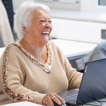 An older white woman with a tan and short white hair is laughing whilst using a laptop.