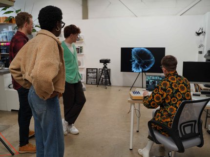 3 people standing in a studio space and looking at a blue abstract image on a screen. 1 person in a sunflower pattern shirt sitting down at a laptop, manipulating the image on screen.