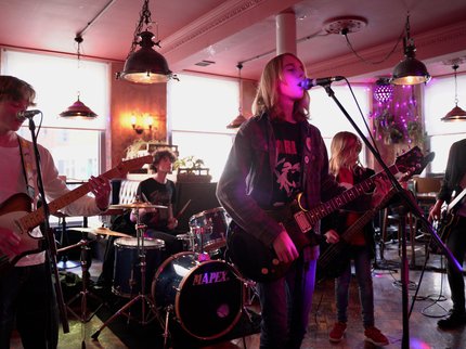 A band playing on stage. 2 people at the front playing guitar and singing into a microphone. 3 people in the background playing guitars and drums.