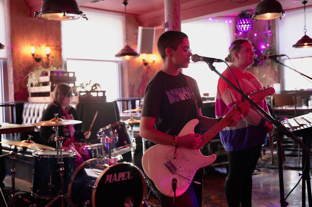 A band playing on stage. 2 people are at the front, one is playing guitar and one is singing into a microphone. There is a person in the background playing drums.