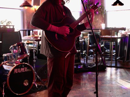 A person wearing a hat, standing on a stage, playing guitar and singing into a microphone