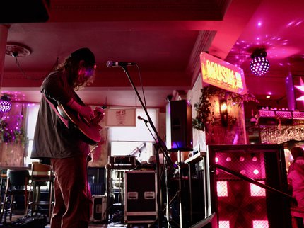 A young person wearing a hat with mid-length hair, playing guitar. They are on a stage with music equipment, lights and a disco ball in the background.