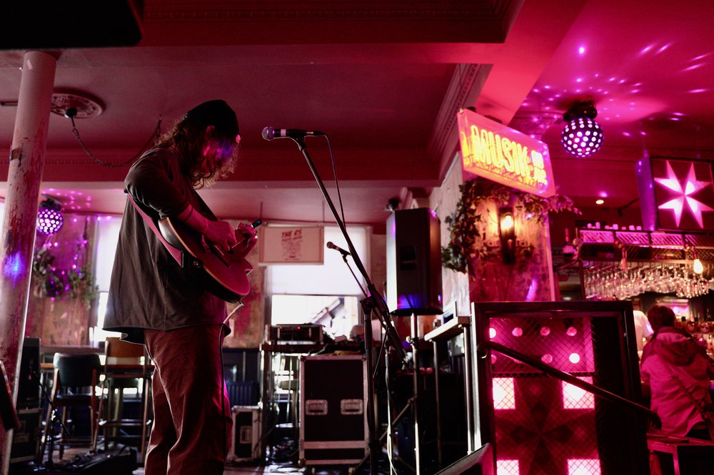 A young person wearing a hat with mid-length hair, playing guitar. They are on a stage with music equipment, lights and a disco ball in the background.