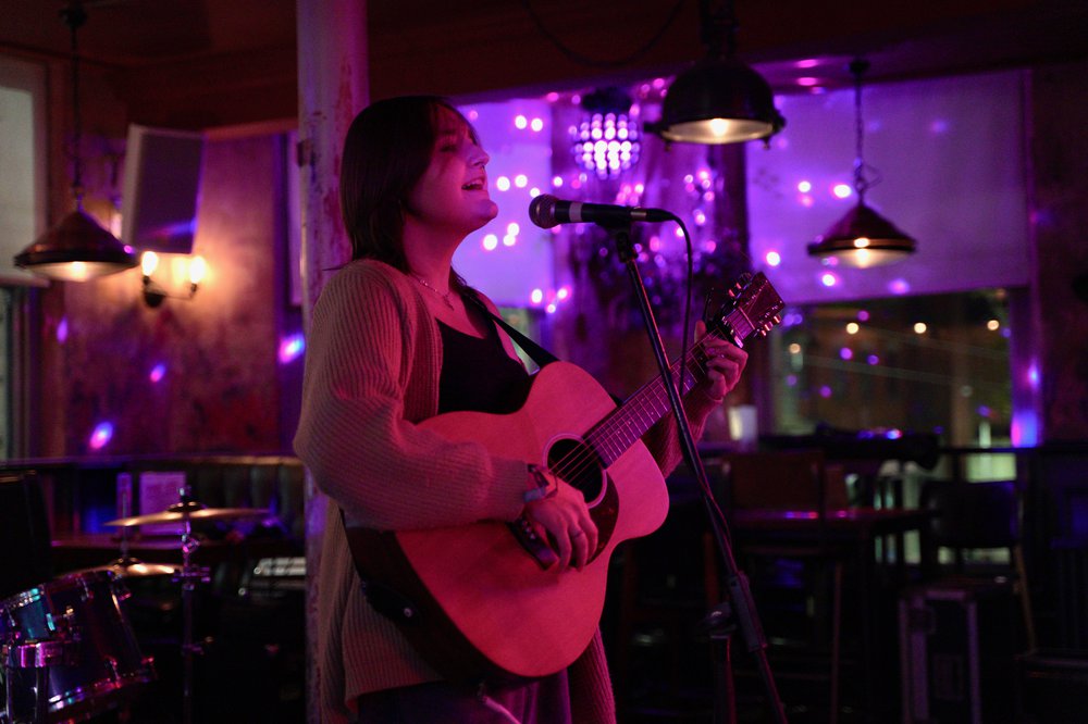 Female singing and holding a guitar. The lights are pink.
