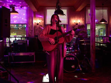 A girl playing acoustic guitar in front of a drum kit