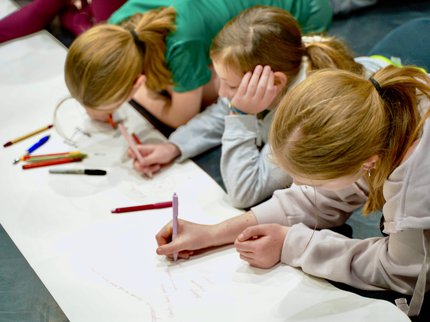 Three young girls drawing on a large piece of paper on the floor.