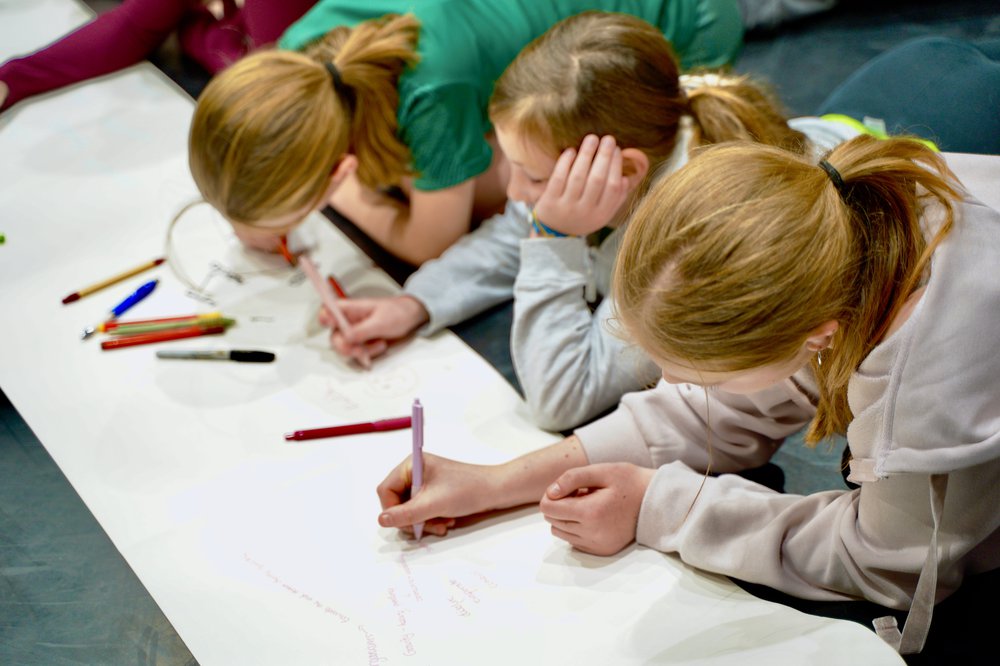 Three young girls drawing on a large piece of paper on the floor.