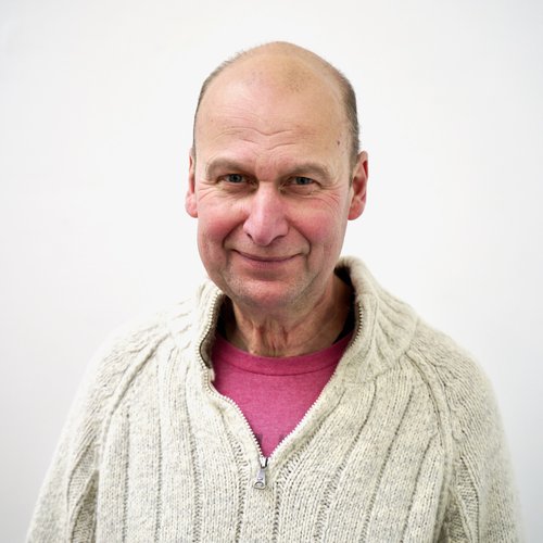 White male, wearing a pink t-shirt under a cream zip down knitted jumper, dark short hair smiling