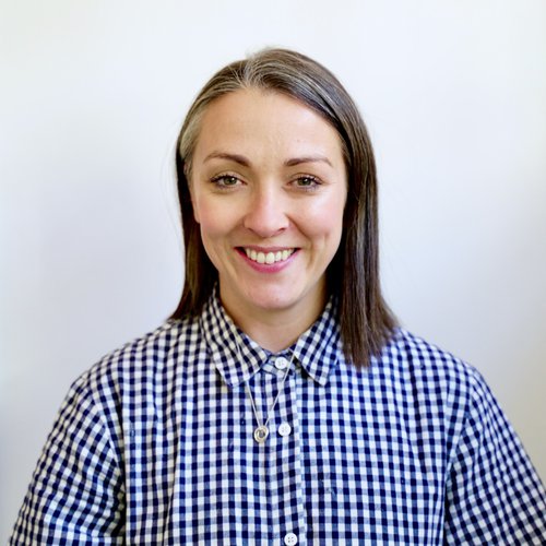 Female with short brown and grey hair, wearing blue and white gingham check shirt, pink lipstick,smiling