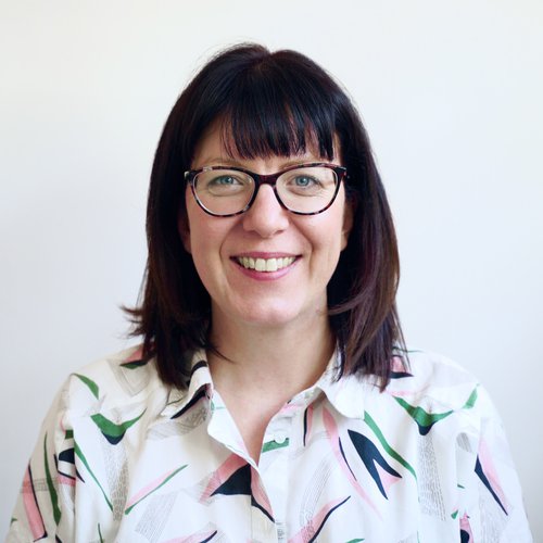 Female, smiling to camera wearing a white shirt with pink, green and blue pattern. Dark brown hair and wearing dark purple framed glasses.
