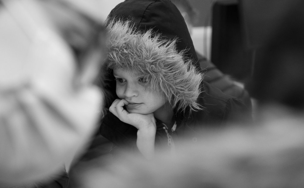 Young boy, in a coat with his hood up, resting on his hand looking down at a table. Image is in black and white