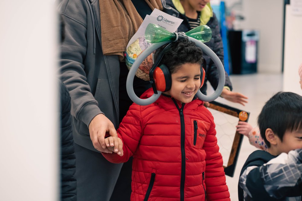 A smiling young boy, in a red coat holding onto a woman's hand, he is wearing large red headphones with tubes coming out the sides.