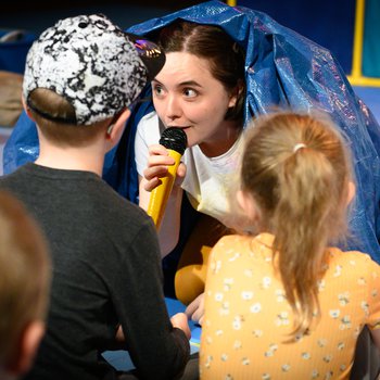 An actor under a sheet of plastic speaks to two children using a microphone.