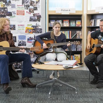 Three guitar players, two female and one male, seated in front of library book shelves.