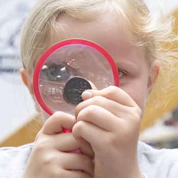A girl with blonde hair holds up an ancient coin to a magnifying glass