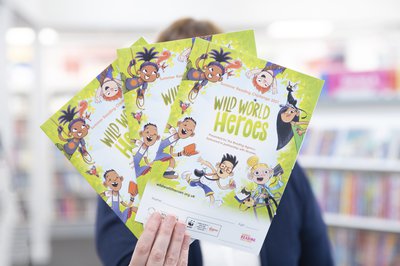 A person, face obscured,  holds up 3 Wild World Heroes collector posters. Library shelves blurred in background.