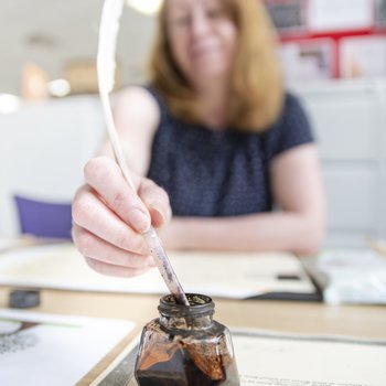 A woman dips a quill pen in a pot of black ink