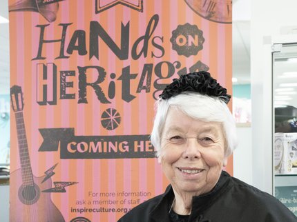 A white-haired lady in Victorian costume, standing in front of the pink and orange striped banner for Hands on Heritage Day