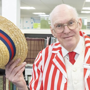 A man wearing glasses and a red and white striped jacket doffs a straw boater toward the camera