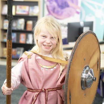A girl with blonde hair, dressed in a pink Viking tunic and holding a spear and shield.