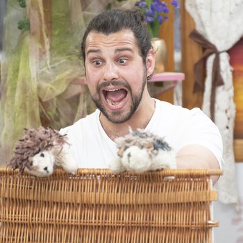 An male actor, looks suprised.  In the foreground two puppet hedgehogs sit on upturned wicker basket lid.