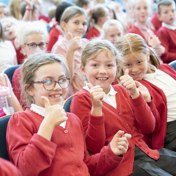 Children in red school jumpers sitting in the audience and smiling with thumbs up