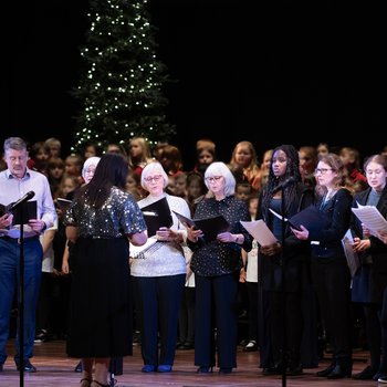 A group of senior choir members sing holding song books, with a christmas tree in the background