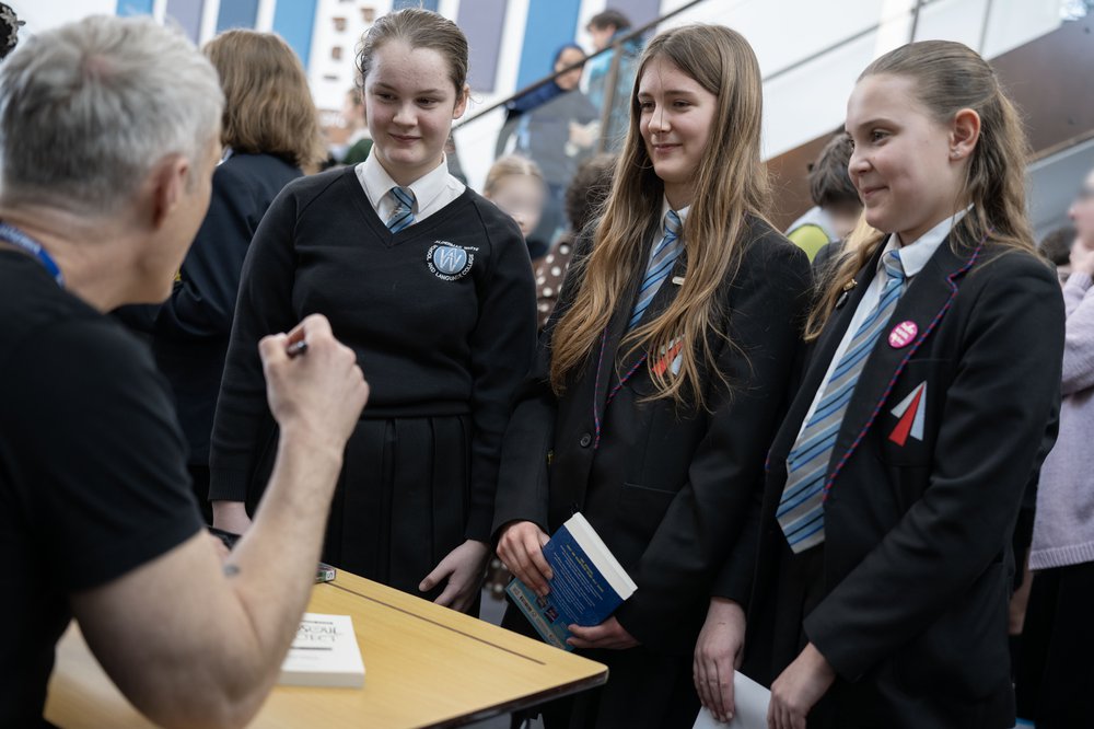 Author Dan Smith sits at a table signing books and three pupils stand