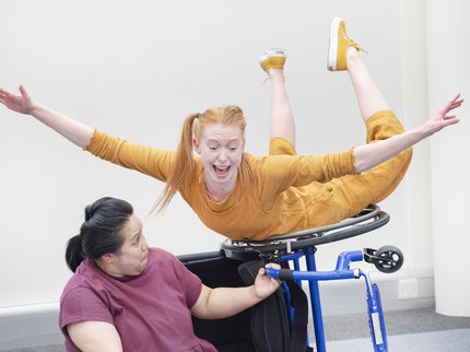 A performer in a wheelchair wearing red clothes supports another performer balance in the air wearing yellow