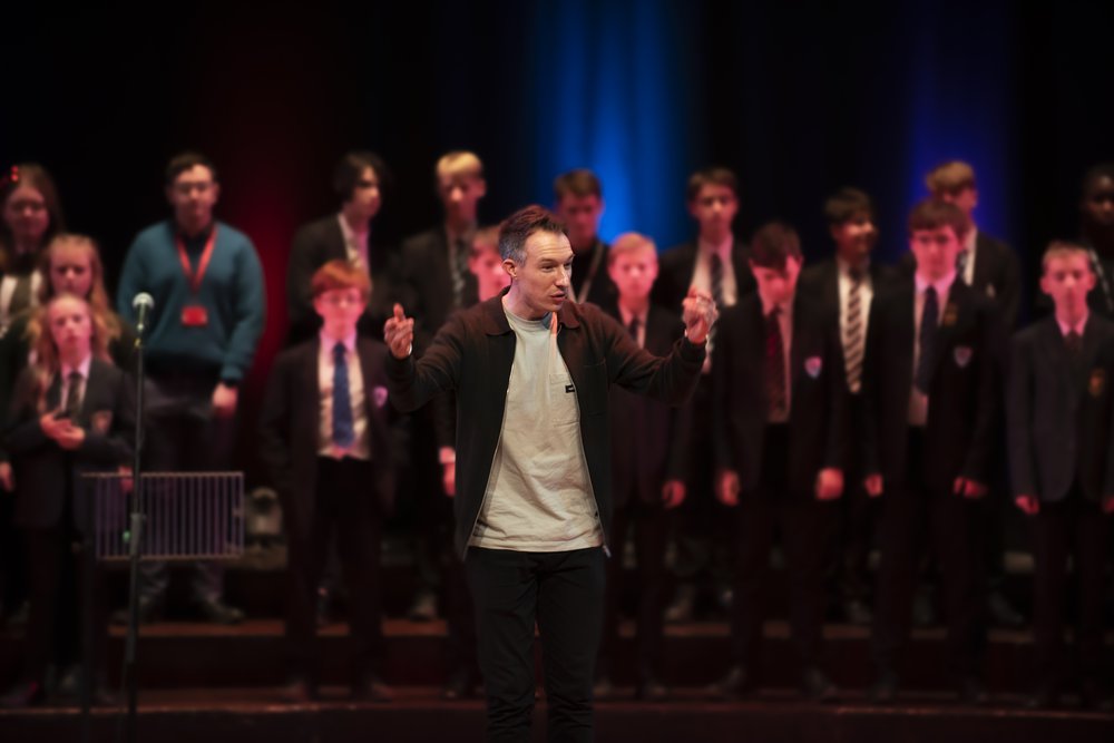 A school group sing on stage with their tutor